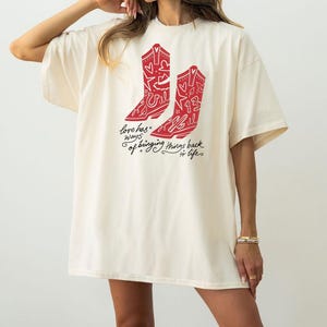 K&ouml;nnte beinhalten: Ein cremefarbenes Kurzarm-T-Shirt mit einer roten Grafik von zwei Cowboystiefeln, die mit Herzen, Sternen und der Zahl 13 verziert sind. Unter den Stiefeln steht in schwarzer Schreibschrift: love has ways of bringing things back to life.
