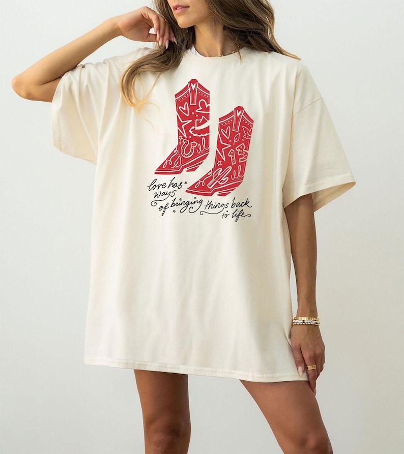 K&ouml;nnte beinhalten: Ein cremefarbenes Kurzarm-T-Shirt mit einer roten Grafik von zwei Cowboystiefeln, die mit Herzen, Sternen und der Zahl 13 verziert sind. Unter den Stiefeln steht in schwarzer Schreibschrift: love has ways of bringing things back to life.