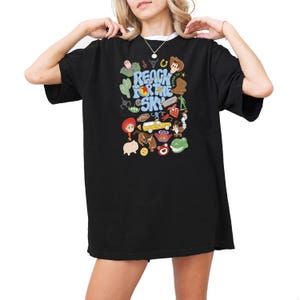 May include: A black short-sleeved t-shirt featuring a colorful graphic print with the text "REACH FOR THE SKY" surrounded by various illustrated characters and icons including a cowboy, space ranger, dinosaur, pig, and a yellow toy car.