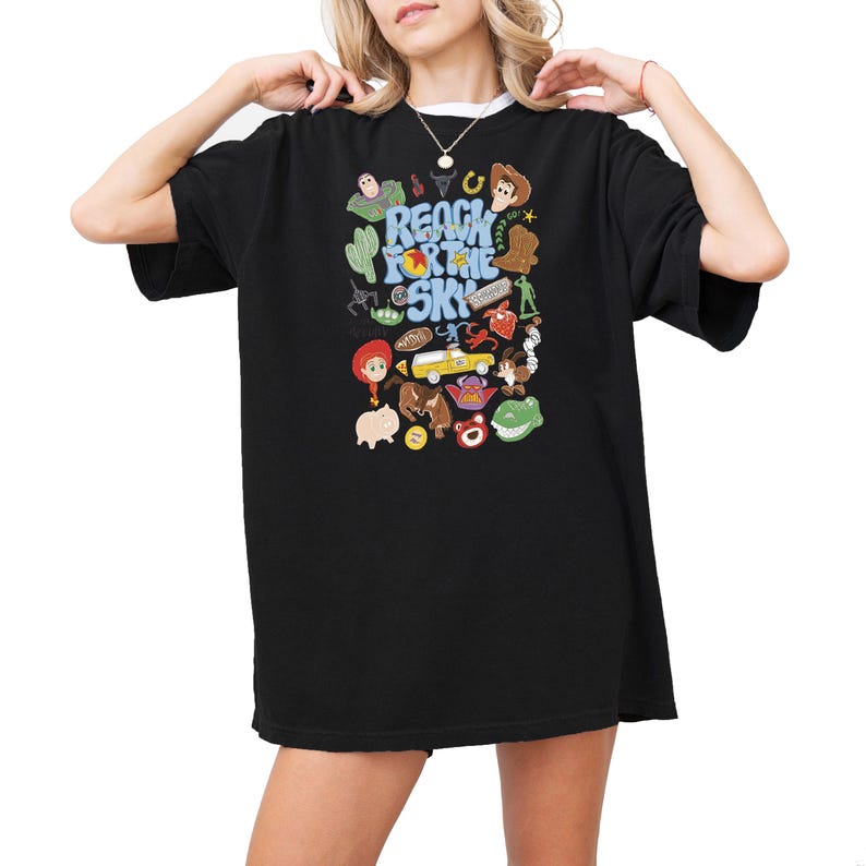 May include: A black short-sleeved t-shirt featuring a colorful graphic print with the text "REACH FOR THE SKY" surrounded by various illustrated characters and icons including a cowboy, space ranger, dinosaur, pig, and a yellow toy car.