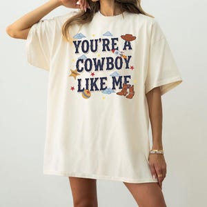 Op de afbeelding: Een off-white t-shirt met korte mouwen en de donkerblauwe tekst "YOU'RE A COWBOY LIKE ME", omringd door kleurrijke grafische illustraties waaronder een bruine cowboyhoed, een raket, een sheriffster, een bal en bruine cowboylaarzen.