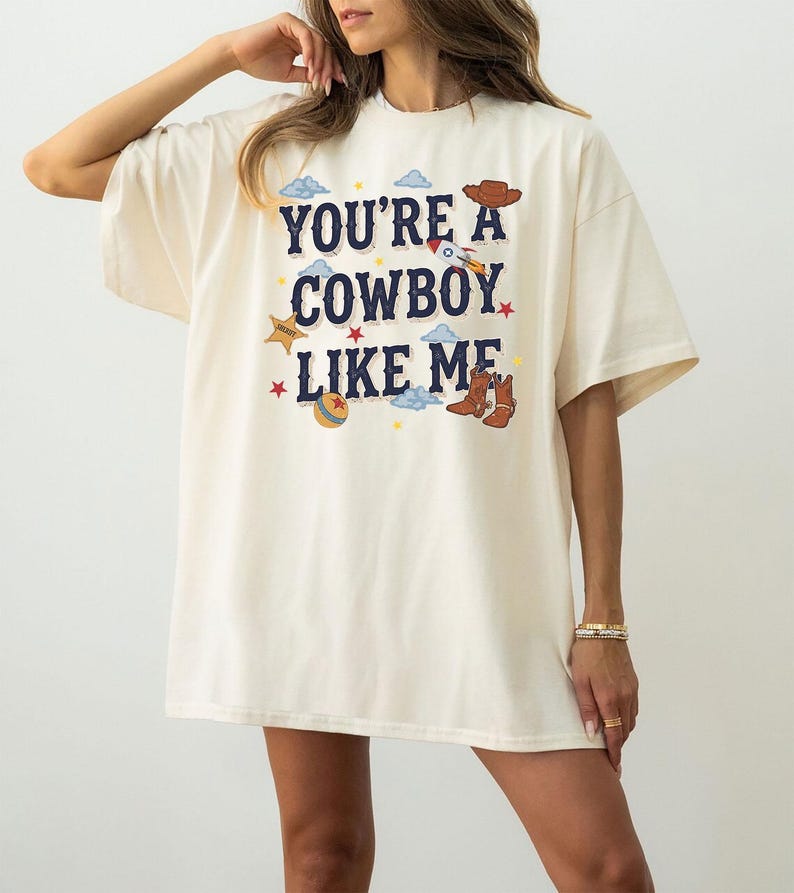 Op de afbeelding: Een off-white t-shirt met korte mouwen en de donkerblauwe tekst "YOU'RE A COWBOY LIKE ME", omringd door kleurrijke grafische illustraties waaronder een bruine cowboyhoed, een raket, een sheriffster, een bal en bruine cowboylaarzen.