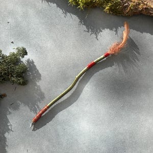 May include: A handcrafted wand made from a natural twig, painted with gold and red accents. The wand features a feather at the tip, adding a whimsical touch. The wand is a unique, decorative item.