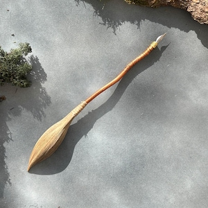 May include: A handcrafted wooden pen with a natural, rustic aesthetic. The pen features a light brown, leaf-shaped nib and a slender, curved wooden body. The handle is wrapped with twine and the pen is made of natural materials.