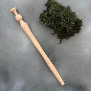 Peut inclure: Un bâton de cheveux en bois clair avec un dessus arrondi et un corps long et effilé. Le bâton de cheveux est posé sur une surface grise à côté d'un touffe de mousse verte. L'accessoire est conçu pour coiffer les cheveux.