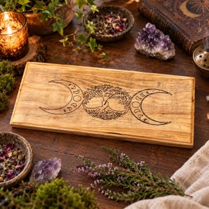 May include: A rectangular wooden board featuring a black laser-engraved design of a central tree of life motif flanked by two crescent moons. The natural light brown wood grain is visible across the flat surface of the board, which has straight edges.