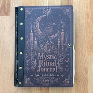 May include: A dark purple journal titled Mystic Ritual Journal with the subtitle Spells Symbols Reflections. The cover features a raven, crescent moon, candles, and crystals in copper ink. A black spine with six brass rivets secures the cover.
