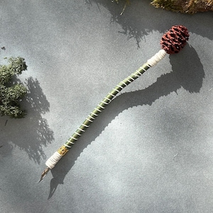May include: A handcrafted calligraphy pen with a natural design. The pen has a green stem bound with white thread, a brown pinecone head, and a metal nib. It is designed for artistic writing and drawing.