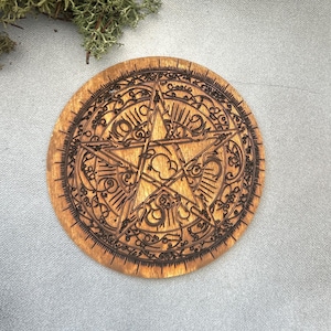 Op de afbeelding: Een ronde houten onderzetter met een gedetailleerd pentagramontwerp. De onderzetter heeft ingewikkelde lasergesneden patronen en een ster in het midden, omringd door sierlijke krullen. Het hout heeft een warme, natuurlijke tint en het ontwerp is donkerbruin.