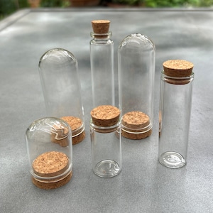 May include: A collection of six clear glass containers of varying heights and shapes, each featuring a removable natural cork stopper. The set includes tall cylindrical vials and rounded dome-shaped glass cloches, all displayed together.