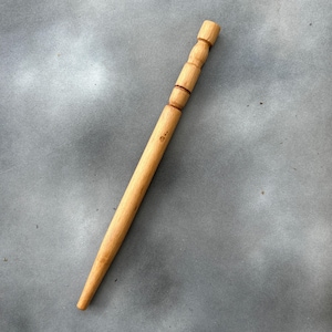 Puede incluir: Una horquilla de madera clara con un diseño cónico. La parte superior presenta una serie de segmentos redondeados y un extremo cilíndrico. La horquilla está diseñada para asegurar el cabello en su lugar.