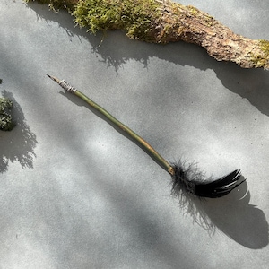 May include: A handcrafted writing quill with a green and gold painted wooden shaft. The quill has silver wire around the nib and a cluster of black feathers. The quill is on a grey surface.