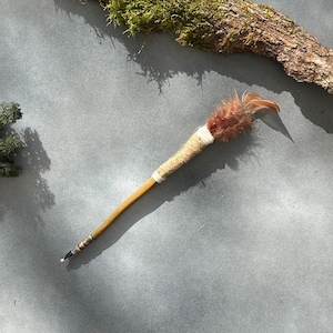 May include: A handcrafted wand with a wooden handle, wrapped in twine and white thread. The wand features a cluster of brown feathers at the top and a metallic tip. The wand is a unique, decorative item.
