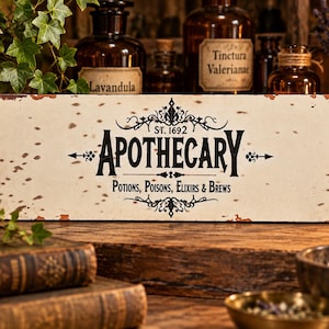 Può includere: Una targa rettangolare in metallo con finitura bianco sporco anticata e bordi color ruggine. Il testo nero recita Apothecary, St. 1692 e Potions, Poisons, Elixirs & Brews, centrato con decorazioni ornamentali e piccole frecce laterali.