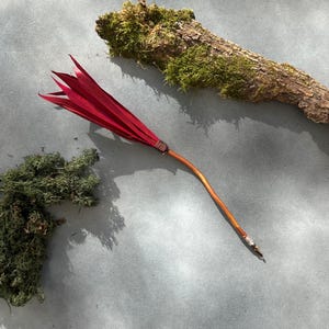 May include: A decorative red and brown broom with a bundle of red leaves at the top. The handle is a light brown colour, and the leaves are tied together with a brown wire. The broom is lying on a grey surface, with moss and a piece of wood in the background.