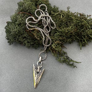 Puede incluir: Collar de metal plateado con un colgante en forma de flecha. El colgante tiene detalles intrincados y una punta dorada. La cadena es de estilo clásico. El collar se muestra sobre un fondo de musgo verde.