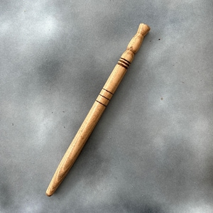 Puede incluir: Una varita mágica de madera hecha a mano con un acabado marrón claro. La varita presenta un diseño cónico con tres bandas marrones oscuras cerca del centro y un mango detallado. La varita mide aproximadamente 30 cm de largo.