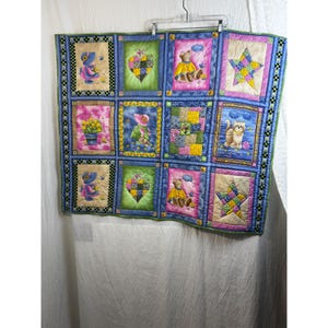 May include: A rectangular patchwork quilt featuring twelve distinct panels with colorful illustrations of teddy bears, cats, hearts, and stars. The quilt is bordered by a green and black checkered trim and hangs from a metal hanger.