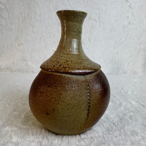May include: A ceramic vase with a rounded body and a narrow neck. The piece features a two-tone glaze with a speckled light olive green top and a darker, textured brown base. A vertical, decorative zigzag stitch pattern runs down the side.
