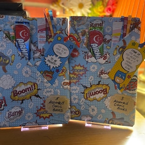 May include: Two rectangular packages wrapped in light blue comic book patterned paper featuring "Boom!" and "Bang!" text. Each contains five assorted comic books, an Airheads candy bar, and a tag with inspirational text. One tag reads "You don't have to be super to be mighty!" and the other "Stand up for what you believe in even if it means standing alone."