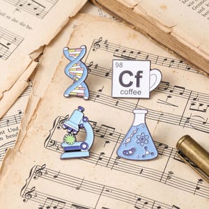 Könnte beinhalten: Vier Emaille-Anstecknadeln mit wissenschaftlichen Motiven: eine hellblaue DNA-Doppelhelix, eine weiße quadratische Kaffeetassen-Ikone mit der Aufschrift 98 Cf coffee, ein hellblaues Mikroskop mit grünem Präparat und ein hellblauer Erlenmeyerkolben.