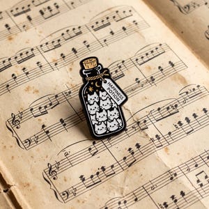 Könnte beinhalten: Eine schwarze Emaille-Anstecknadel in Form einer Glasflasche, gefüllt mit weißen Katzengesichtern. Die Flasche hat einen Korken, eine Kordel und ein weißes Etikett mit dem Text HAPPINESS POTION. Goldene Sterne und eine Mondsichel sind oben zu sehen.