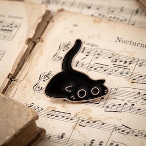 Könnte beinhalten: Eine schwarze Emaille-Anstecknadel in Form einer Katzensilhouette mit erhobenem Schwanz. Die Katze hat goldfarbene Metallumrandungen und goldfarbene Metalldetails, die den Text "COO" bilden, mit kleinen Schnurrhaaren auf der rechten Seite.