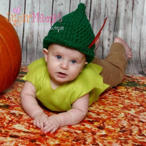 May include: A baby wearing a green crochet hat with a red feather, a lime green top, and brown pants. The baby is lying on a colorful patterned surface. The hat has a pointed top. The text "Sugar Mamas" is visible in the upper left corner.