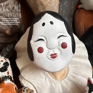 May include: A hand-carved wooden mask with a white face, black hair, red cheeks, and a red smiling mouth. The mask is part of a collection of wood Christmas ornaments. The text "Wood Carving" is at the top.