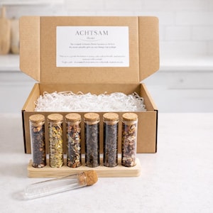 Puede incluir: Un juego de regalo de té con seis viales de vidrio, cada uno con un tapón de corcho, llenos de diferentes tés. Los viales se colocan en un soporte de madera dentro de una caja de cartón abierta con relleno blanco. La caja tiene una etiqueta que dice "ACHTSAM Home."