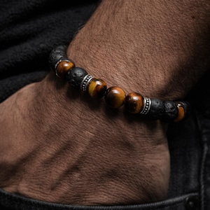 Könnte beinhalten: Ein Armband mit schwarzen Lavastein- und Tigeraugen-Perlen. Das Armband hat abwechselnd schwarze und braune Perlen mit silberfarbenen Abstandshaltern. Das Armband wird am Handgelenk getragen, mit dunklem Hintergrund.