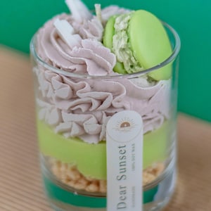 Puede incluir: Una vela con temática de postre en un vaso transparente. La vela presenta capas de cera verde claro y malva, rematadas con un macaron verde y decoraciones blancas. Una etiqueta dice "Dear Sunset".