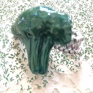 Puede incluir: Un anillo de cerámica verde con un diseño de cuentas plateadas y moradas. El anillo tiene forma de capullo de flor.