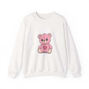 May include: White sweatshirt featuring a pink teddy bear illustration. The bear has a heart on its chest and is in a sitting position. The sweatshirt has a crew neck and long sleeves. The teddy bear design is centered on the front of the garment.