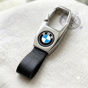 May include: A silver-toned metal keychain featuring a spring-loaded carabiner clip and a black leather loop. The central rectangular metal plate displays the circular BMW logo with its signature blue, white, and black color scheme.