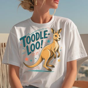 Op de afbeelding: Wit T-shirt met een cartoon kangoeroe en de tekst "TOODLE-LOO!" in turquoise en geel. De kangoeroe is bruin en staat rechtop. Het ontwerp bevat kleurrijke sterren en lijnen rond de tekst en de kangoeroe.