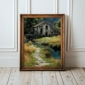 May include: A framed landscape painting depicting a stone cottage situated beside a small stream and a stone bridge. The scene features a winding stone path leading through tall green and golden grasses toward the building, all enclosed in a brown wooden frame.