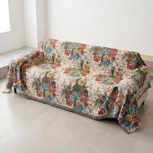 May include: A sofa cover with a floral design. The cover has a cream base, adorned with a pattern of red, blue, yellow, and green flowers. It has a grey fringed edge and is draped over a white sofa.