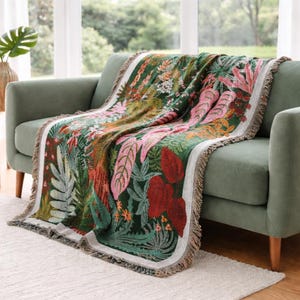 May include: A woven throw blanket with a botanical design featuring large pink, red, and green leaves and flowers. The blanket has a white border and fringed edges. It is draped over a green sofa in a well-lit room.