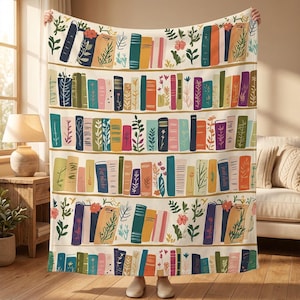 Pode incluir: Uma manta creme com um design colorido de estante. O design inclui filas de livros em várias cores, com detalhes florais e botânicos. A manta é segurada por uma pessoa.