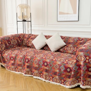 Puede incluir: Un sofá cubierto con una manta estampada en tonos rojos, naranjas y azules. El diseño geométrico presenta formas de diamantes y rectangulares. Dos cojines cuadrados blancos descansan sobre el sofá. La manta tiene un borde con flecos.