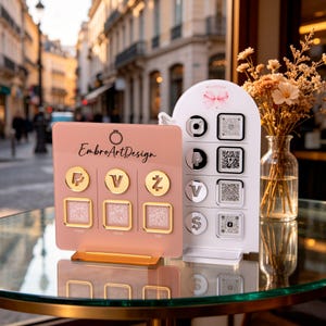 Puede incluir: Dos expositores con códigos QR y símbolos. Uno es rosa con detalles dorados y el texto "EmbroArtDesign". El otro es blanco con un logotipo de lazo rosa. También se ve un jarrón con flores secas.