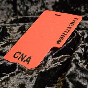 May include: A bright orange rectangular tag with the text "THEY/THEM" and "CNA" in black. The tag has a small notch at the bottom and a slit at the top. The background is a dark, textured fabric.