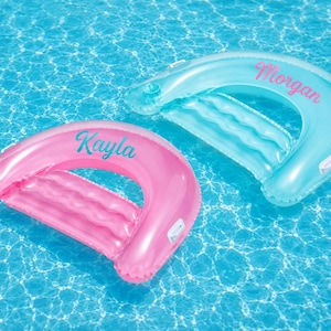 May include: Two inflatable pool floats in a U-shape design, one translucent pink with the name Kayla in teal script and one translucent light blue with the name Morgan in pink script, each featuring a built-in cup holder and a ribbed backrest.