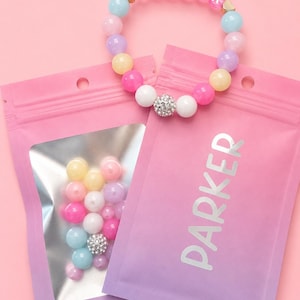 May include: A beaded bracelet featuring pastel-colored round beads, a central rhinestone-encrusted bead, and letter beads spelling PARKER. Two pink gradient pouches are included, one with a clear window showing loose beads and one labeled PARKER.