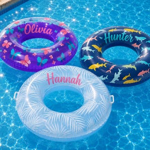 Op de afbeelding: Drie ronde opblaasbare zwembanden drijvend op het water. Een paarse zwemband heeft kleurrijke vlinders en de naam Olivia. Een donkerblauwe zwemband heeft diverse haaien en de naam Hunter. Een witte zwemband heeft palmbladeren en de naam Hannah.