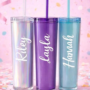May include: Three personalized tumblers in iridescent, purple, and teal, each with a matching lid and straw. The names "Riley," "Layla," and "Hannah" are written in white script on each tumbler. The background is pink with confetti.