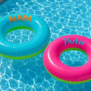 May include: Two inflatable pool rings floating on water. One ring is bright blue with the name ALANA printed in orange. The other ring is hot pink with the name Parker printed in blue. A small multi-coloured beach ball floats nearby in the water.