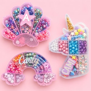 Puede incluir: Tres recipientes de almacenamiento de plástico transparente con forma de concha, arcoíris y unicornio, llenos de cuentas de colores variados. La concha tiene una estrella rosa con la letra A, el arcoíris el nombre Carly y el unicornio CAMI.