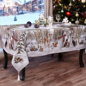 May include: A rectangular tablecloth featuring a winter forest scene with snow-covered birch trees, a snowman, a deer, and various woodland animals. The fabric has a light grey background with white tassels hanging from each of the four corners.